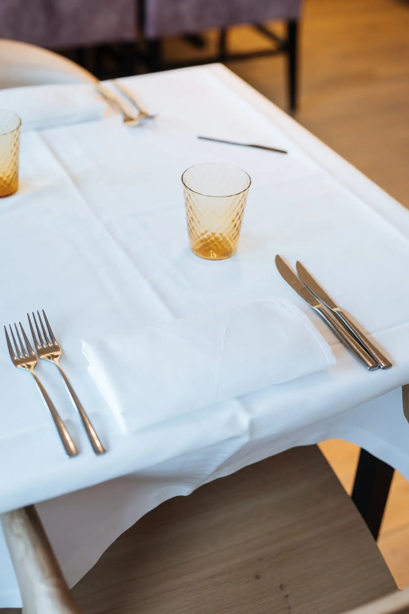 Een gedekte tafel met wit linnen, bestek en een amberkleurig glas